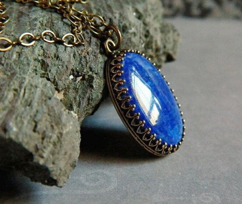 May include: An oval-shaped blue lapis lazuli gemstone pendant set in a decorative bronze-toned metal bezel. The pendant hangs from a matching bronze-toned metal chain, featuring a smooth, polished surface that reflects light across its deep blue hue.