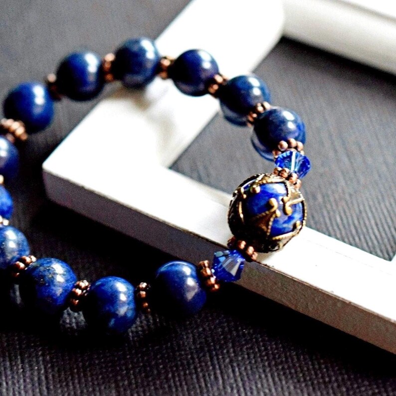 A blue lapis lazuli beaded bracelet with copper spacer beads and a decorative metal charm. The charm is round and has a blue lapis lazuli stone in the center. The bracelet is on a white surface.