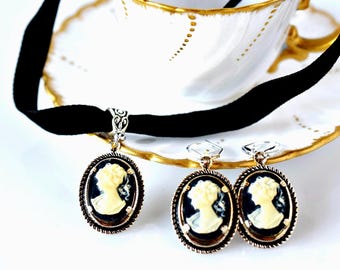 May include: A black velvet choker necklace and a pair of matching drop earrings, each featuring an oval cameo pendant with a cream-colored profile silhouette of a person against a dark background, set in a textured silver-toned metal frame.