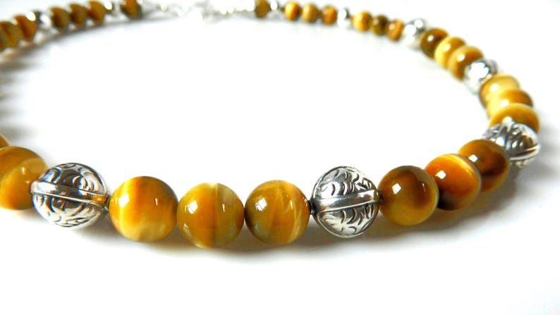 May include: A beaded necklace featuring round, polished golden-brown tiger's eye gemstones alternating with spherical, silver-toned metal beads that have an intricate, engraved leaf-like pattern throughout the surface of the metal.