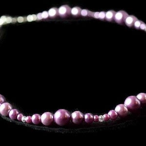 Puede incluir: Un collar con una hebra de cuentas redondas moradas de diferentes tama&ntilde;os. Las cuentas est&aacute;n ensartadas, formando una forma circular. Peque&ntilde;as cuentas de acento transparentes est&aacute;n intercaladas en el collar. El fondo es negro liso.