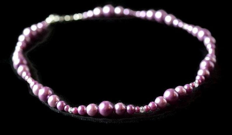 Puede incluir: Un collar con una hebra de cuentas redondas moradas de diferentes tama&ntilde;os. Las cuentas est&aacute;n ensartadas, formando una forma circular. Peque&ntilde;as cuentas de acento transparentes est&aacute;n intercaladas en el collar. El fondo es negro liso.