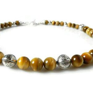 May include: A necklace featuring round, golden-brown tiger's eye beads alternating with polished, etched silver-toned metal beads. The beads are arranged in a circular pattern, connected by a silver-toned clasp at the back of the jewelry piece.