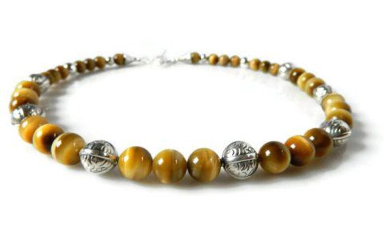 May include: A necklace featuring round, golden-brown tiger's eye beads alternating with polished, etched silver-toned metal beads. The beads are arranged in a circular pattern, connected by a silver-toned clasp at the back of the jewelry piece.