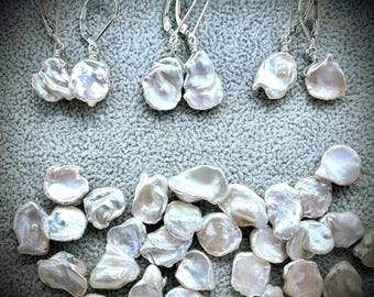 May include: Three pairs of silver leverback earrings featuring irregular, iridescent white baroque pearls, accompanied by a collection of loose, matching baroque pearls scattered below the earrings on a textured gray surface.