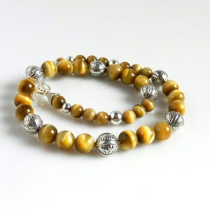 May include: A beaded necklace featuring round, golden-brown tiger's eye gemstones interspersed with polished silver-tone metal beads. The silver beads alternate between smooth, spherical shapes and larger, textured spheres with intricate floral engravings.