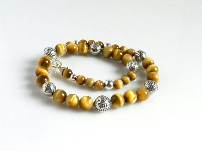 May include: A beaded necklace featuring round, golden-brown tiger's eye gemstones interspersed with polished silver-tone metal beads. The silver beads alternate between smooth, spherical shapes and larger, textured spheres with intricate floral engravings.