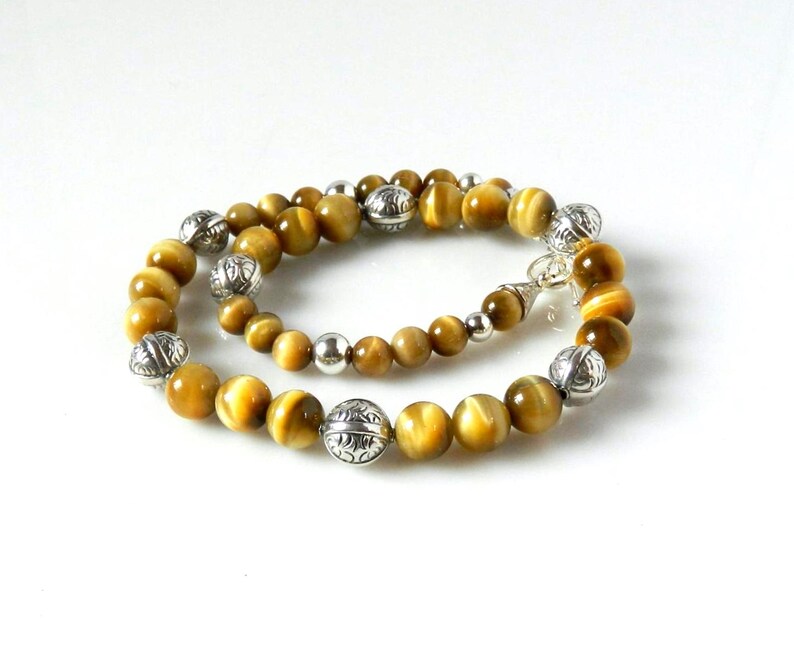 May include: A beaded necklace featuring round, golden-brown tiger's eye gemstones interspersed with polished silver-toned metal beads. The necklace includes ornate, etched silver-toned spherical beads and smaller, smooth silver-toned spacer beads.