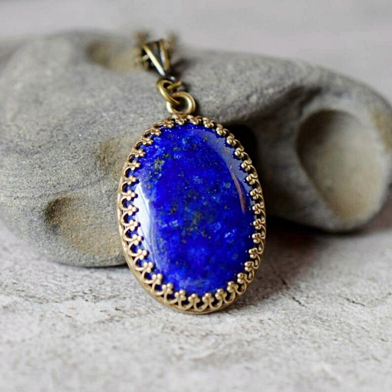 May include: An oval pendant necklace featuring a deep blue lapis lazuli stone with natural gold-colored flecks. The stone is set in a decorative, scalloped antique brass-toned metal frame with a matching metal chain loop at the top.