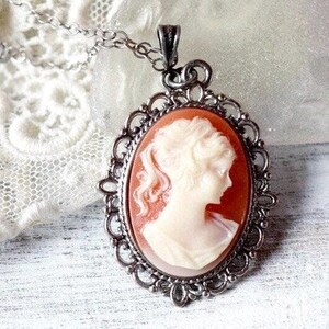 May include: A silver cameo necklace with a delicate filigree design. The cameo features a white profile of a woman against a peach background.