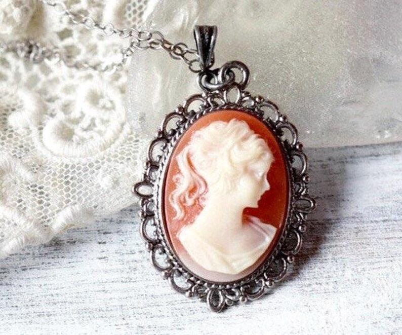 May include: A silver cameo necklace with a delicate filigree design. The cameo features a white profile of a woman against a peach background.