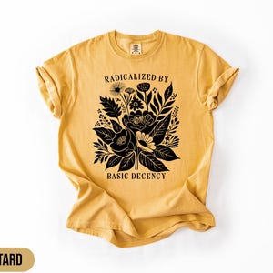 Könnte beinhalten: Senffarbenes T-Shirt mit schwarzem Blumendesign und dem Text "RADICALIZED BY BASIC DECENCY". Die Grafik zeigt verschiedene Blumen und Blätter. Das Shirt hat kurze Ärmel und einen Rundhalsausschnitt.