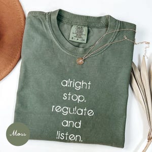 May include: A moss green t-shirt with the words "alright stop, regulate and listen" printed in white. A gold necklace with a pendant rests on the shirt. A brown hat and dried leaves are in the background.