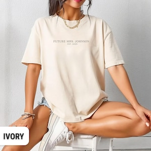 May include: Ivory-coloured t-shirt with the text "FUTURE MRS. JOHNSON EST. 2025" printed in a light brown colour. The shirt is a relaxed fit and has short sleeves. The model is wearing denim shorts and white trainers.