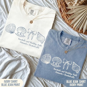 Könnte beinhalten: Zwei T-Shirts, eines in Elfenbein und eines in Jeansblau, mit Muschel- und Schleifenmotiven. Der Text lautet "Seashells and Wedding Bells Amanda's Bachelorette". Jedes Shirt hat eine goldene Halskette. Das elfenbeinfarbene Shirt hat einen Jeansdruck.