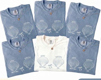 May include: A collection of blue jean and ivory t-shirts with a seashell design. Each shirt features the text "Olivia's Beach Bach Monte Carlo 2026" or "2025" in white. The shirts are adorned with a gold-colored necklace.