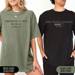May include: Two t-shirts, one moss green and one black, with pregnancy announcement text. The moss green shirt says "Growing a Tiny Human" with "May 2026". The black shirt says "Dad Mode Loading" with "Est. 2026".