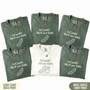 May include: Six t-shirts, five in moss green and one in ivory, are displayed. The shirts have the phrase "last pickle She'll ever tickle" with a pickle graphic, except one that says "I'll ever tickle" with a ring graphic. Each shirt is adorned with a necklace.