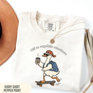 Puede incluir: Camiseta de color marfil con un dibujo de un ganso en monopatín, con gorra y gafas de sol, sosteniendo una taza de café. El texto "Off to regulate emotions" está sobre el dibujo. También se ven un collar dorado y un sombrero marrón.
