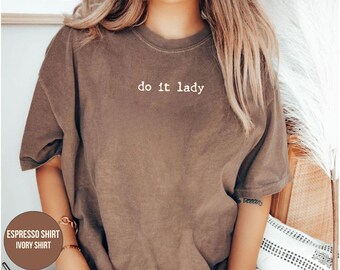 Puede incluir: Camiseta color espresso con el texto blanco "do it lady". La camiseta es de estilo casual de manga corta. La imagen también muestra un vaso de café helado y una pulsera.