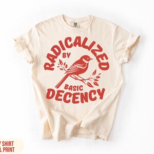 May include: Ivory t-shirt with a coral-coloured graphic. The graphic shows a bird on a branch, with the words "RADICALIZED BY BASIC DECENCY" in a semi-circular arrangement above and below the bird.