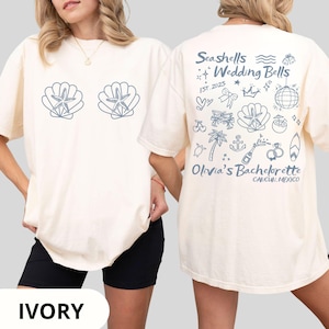 May include: Ivory-colored t-shirt with a seashell and starfish design on the front. The back features the text "Seashells & Wedding Bells" with various beach-themed illustrations and "Olivia's Bachelorette Cancun, Mexico."