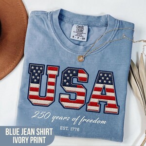 Puede incluir: Una camiseta Comfort Colors azul claro con la palabra "USA" bordada en rojo, blanco y azul. La camiseta presenta el texto "250 years of freedom" y "EST. 1776". Un collar de oro descansa sobre la camiseta. La camiseta está etiquetada como "BLUE JEAN SHIRT IVORY PRINT".