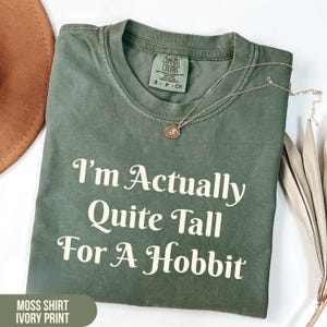 May include: A moss green t-shirt featuring the ivory text I'm Actually Quite Tall For A Hobbit printed on the front. The shirt has a crew neckline and a Comfort Colors brand label inside the collar, accompanied by a thin gold-toned necklace.
