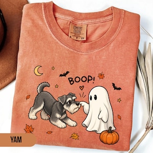 Puede incluir: Una camiseta de cuello redondo color naranja quemado con un gráfico de un perro schnauzer gris y blanco interactuando con un fantasma blanco. El texto "BOOP!" está impreso encima, rodeado de pequeños murciélagos, estrellas, hojas de otoño y una calabaza.