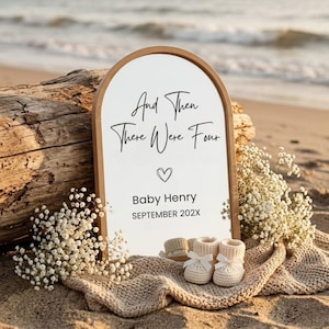 Puede incluir: Un letrero arqueado con marco de madera clara que presenta el texto negro "And Then There Were Four", un icono de corazón, "Baby Henry" y "SEPTEMBER 202X". Dos pequeños patucos de bebé de punto color crema con lazos blancos están delante.
