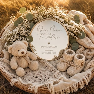Peut inclure: Un miroir rond avec le texte One More To Adore, BABY ANDRESON, ARRIVING SEPTEMBER 2026, placé dans un panier en osier avec un ours en peluche beige, des chaussons tricotés, des fleurs blanches et un tissu en dentelle crème.