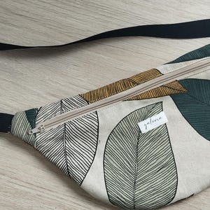 May include: A beige fanny pack with a black adjustable strap. The pack has a zipper closure and a green and brown leaf pattern. The word "galena" is printed on the front of the pack.