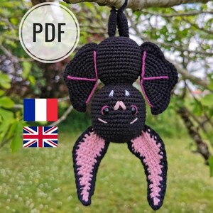 Könnte beinhalten: Eine schwarze gehäkelte Fledermaus mit rosa Flügeln, die an einem Ast hängt. Die Fledermaus hat ein süßes Gesicht mit weißen Augen und einer rosa Nase.