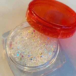 Puede incluir: Un recipiente cuadrado transparente lleno de purpurina iridiscente. El recipiente tiene una base transparente y una tapa naranja translúcida abierta. La purpurina brilla con varios colores, incluyendo azul, verde y amarillo.