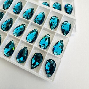 May include: Teardrop-shaped teal crystal rhinestones in a white plastic tray. Each gem has a faceted surface and a small hole for stringing. The tray is divided into individual compartments. The gems are a vibrant shade of teal.