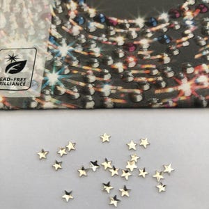 May include: A collection of small, gold-colored star-shaped embellishments scattered on a white surface. The word "PRECIOSA" is visible on a partially visible package in the background. The package also has a "LEAD-FREE BRILLIANCE" logo.