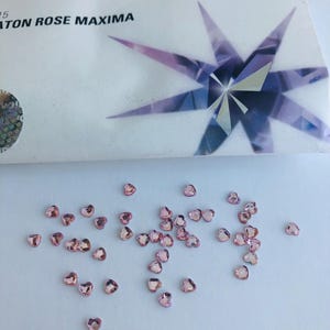 Könnte beinhalten: Kleine, herzförmige, rosa Strasssteine, die auf einer weißen Oberfläche verstreut sind. Die Strasssteine befinden sich in der Nähe einer weißen Schachtel mit dem Wort "PRECIOSA" und dem Text "CHATON ROSE MAXIMA" sowie einem Sternmuster.