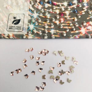Op de afbeelding: Kleine, vlindervormige versieringen in roségoud en zilver tinten liggen verspreid op een witte ondergrond. De afbeelding toont ook een deel van een verpakking met het woord "PRECIOSA" en een "LEAD-FREE BRILLIANCE" logo.