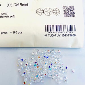 Puede incluir: Un montón de cuentas XILION transparentes e iridiscentes con un paquete en el fondo. El paquete tiene la palabra "SWAROVSKI", el texto "XILION Bead" y un código de barras. Las cuentas están dispersas sobre una superficie blanca.