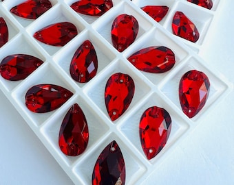 May include: A white tray filled with various shapes of faceted, deep red rhinestones. The rhinestones include teardrop, oval, and irregular shapes, each with a hole for attachment. The tray's grid pattern neatly organizes the sparkling gems.