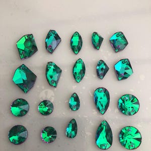Puede incluir: Un surtido de pedrería verde esmeralda en varias formas, incluyendo redondas, de lágrima y diseños geométricos. La pedrería tiene una calidad reflectante e iridiscente, y está dispuesta sobre una superficie clara. Estos adornos decorativos son adecuados para manualidades y la fabricación de joyas.
