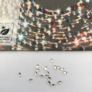 May include: A scattering of small, oval-shaped, clear rhinestones. The rhinestones are scattered on a white surface. The word "PRECIOSA" is visible in the background, along with a design of colorful rhinestones.