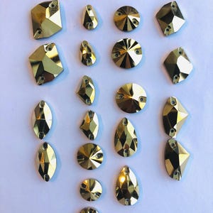 May include: A collection of various gold-colored rhinestones in different shapes, including round, teardrop, and geometric designs. Each rhinestone has a faceted surface for enhanced sparkle and shine. The rhinestones are arranged on a white surface.