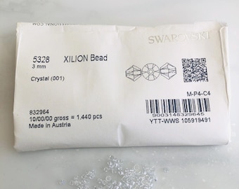 May include: A package of Swarovski XILION beads, 3 mm in size, with the text "Crystal (001)". A small pile of clear, faceted beads is scattered below the package. The package includes a barcode and the text "Made in Austria".