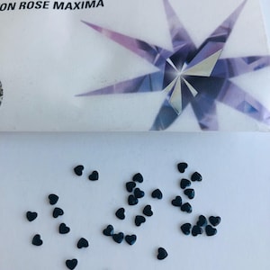 Könnte beinhalten: Kleine, dunkle herzförmige Verzierungen, die auf einer weißen Oberfläche verstreut sind. Das Bild zeigt auch eine Verpackung mit den aufgedruckten Wörtern "PRECIOSA" und "ON ROSE MAXIMA" sowie ein Sternmuster.