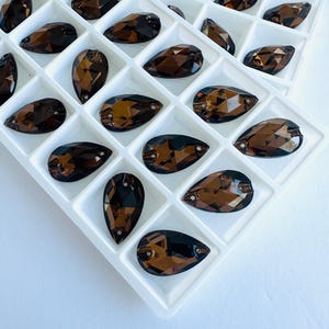 May include: Brown teardrop-shaped rhinestones displayed in a white plastic organiser. The faceted surfaces of the rhinestones reflect light. The rhinestones are a dark, transparent brown colour.