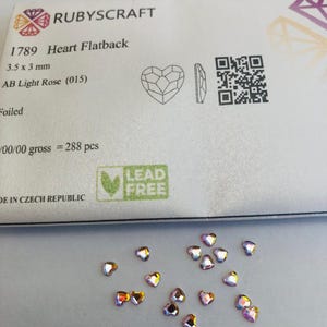Puede incluir: Un conjunto de pedrería plana en forma de corazón iridiscente, de aproximadamente 3,5 x 3 mm, dispersa sobre una superficie blanca. El embalaje dice "RUBYSCRAFT" y "1789 Heart Flatback". La pedrería tiene un acabado AB Light Rose.