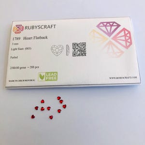Op de afbeelding: Een verpakking hartvormige flatback strass-steentjes in de kleur Light Siam (003), 3 mm groot. De verpakking bevat 288 stuks en is voorzien van de tekst "RUBYSCRAFT" en "LEAD FREE". Verspreid liggen losse rode hartvormige strass-steentjes.