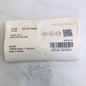 May include: A packet of Swarovski XILION beads, 2.5 mm in size, labelled "Crystal Aurore Boreale (AB)". A small pile of iridescent crystal beads is scattered below the packet. The packet includes a barcode and the text "Made in Austria".