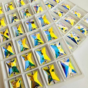 May include: Rectangular, iridescent crystal rhinestones in a white tray. Each crystal is faceted, displaying yellow, blue, and green hues. The crystals are arranged in individual compartments, ready for crafting or embellishment.